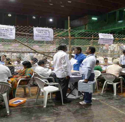 एक टेबल पर रखे मतपत्र और चल रही मतगणना प्रक्रिया, जो चुनाव पारदर्शिता को दर्शाती है