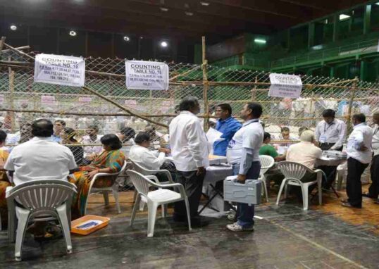 एक टेबल पर रखे मतपत्र और चल रही मतगणना प्रक्रिया, जो चुनाव पारदर्शिता को दर्शाती है