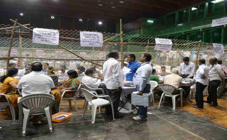 एक टेबल पर रखे मतपत्र और चल रही मतगणना प्रक्रिया, जो चुनाव पारदर्शिता को दर्शाती है